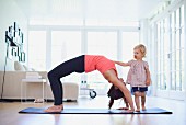 Frau macht Gymnastikübung im Wohnzimmer, daneben die neugierige Tochter
