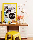 Pin board in yellow-painted antique frame and vintage ornaments on 70s desk next to yellow wooden stool
