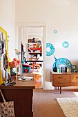 Floral wall stickers above 70s sideboard, yellow-framed pin board above desk and doorway leading into a colourful room