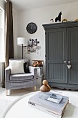 Corner of comfortable living room with pale grey armchair, collage on wall and farmhouse cupboard painted dark grey