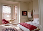 Double bed with red counterpane, seating area in window bay and pattern of light and shade falling through closed louvre blinds