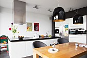 Dining table below black pendant lamps in white, open-plan kitchen with free-standing counter and fitted appliances