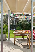 Tisch auf überdachter Terrasse mit buntem Girlandenschmuck und Blick in den Garten