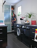 Modern laundry room with washing machine integrated into black base cabinets; woman in front of cupboard in background