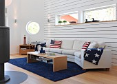 White sofa combination with stars and stripes cushions and blankets below ribbon window in wood-clad wall and porthole in background