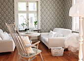 Gemütliches Wohnzimmer - weiße Sofagarnitur und Schaukelstuhl um Tisch am Fenster, an Wand gemusterte Tapete