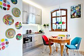 Brightly coloured, retro, upholstered chairs around antique, exotic-wood table, stainless steel kitchen counter and wall plates in open-plan kitchen