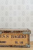 Old wooden crate with black stencilled lettering against wallpapered wall with pale grey retro pattern