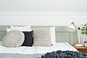 Verschiedene Kissen in Weiß- und Grautönen auf Bett, an grau lackiertem Holz Kopfteil