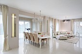Luxuriöser Wohnraum in Weiß, Essplatz mit postmodernem Tisch und gepolsterten Stühle, im Hintergrund Sofalandschaft auf hochglänzendem Fliesenboden