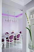 Dining table and crystal chandelier in dining room with purple, indirect lighting on ceiling and Neo-Baroque, upholstered chairs