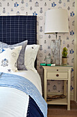 Bed with blue headboard and country-style wallpaper, bedside table with lamp