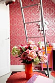 Bouquet in orange glass vase on doily in front of metal ladder and pink, patterned wallpaper