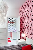 Bed against wall with shoe-patterned, pink wallpaper, view into ensuite bathroom with black polka-dot wall