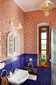Corner of vintage bathroom with blue-painted wainscoting, patterned wallpaper, paraffin-style lamp and narrow window