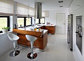 Designer bar stools at counter under cylindrical, stainless steel extractor hood in open-plan kitchen