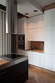 Island counter with integrated hob below cylindrical, stainless steel extractor hood next to fitted kitchen units with flat, white fronts