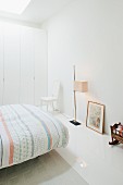 Bed with patterned bedspread, standard lamp and fitted wardrobes in minimalist, white bedroom