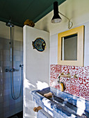 Trough-style sink below wall tiles with red and white pattern of circles and shower area behind partition in rustic bathroom