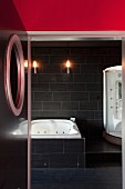 Futuristisches Badezimmer, schwarz gefliest mit Whirlpool-Wanne und moderner Duschkabine; Blick durch geöffnete Tür mit Bullauge