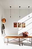 Pattern of light and shade in hallway with bulb pendant lamps above wooden bench, artworks on wall and coat hooks to one side with original storage bin