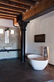 Free-standing designer bathtub with floor-mounted taps on stone-tiled floor in front of black metal column