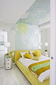 Blick durch offene Tür auf Doppelbett mit Bettwäsche in weissen und gelben Farbtönen, an Wand und Decke Tapete mit Blumenmuster