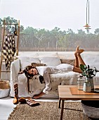 Young woman lying on white loose-covered sofa with fur blanket, simple oak coffee table and picture of woodland projected on wall in harmonious interior in natural shades