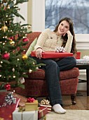 Young woman holding Christmas present sitting next to Christmas tree