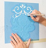 Selbstgemachte Ornamentschablone mit hellblauer Farbe ausgemalt; Hände mit Malerpinsel halten die Schablone fest