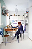 Young woman sitting at kitchen table below cooking utensils hanging from shelf on wall in front of white fitted kitchen