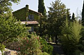 Berankte Giebelseite eines umgenutzten, historischen Gebäudes mit Natursteinmauer, alten Bäumen und Oleander im umgebenden Garten