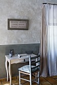 Framed embroidery above small, antique desk with rush-bottomed chair; marbled walls and curtains in natural shades