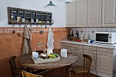 Mediterranean country-house kitchen counter with grey cupboards and white tiles, dining table, wooden chairs, and chalkboard calender above terracotta dado
