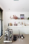 Chrome espresso machine next to sink on counter in front of crockery on wall-mounted shelves