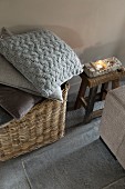 Stack of cushions with grey and brown covers in basket and narrow wooden stool on grey tiled floor