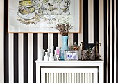 Vases and ethnic artworks on white radiator cover against wallpaper with wide black and white stripes