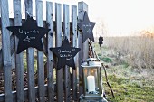 Star-shaped signs made from black-painted wooden hanging on a fence with a lantern with a candle with someone walking in the background