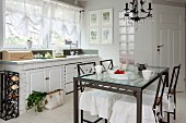 Dining table with glass top and metal frame, chairs with white loose covers and kitchen counter below window with translucent gathered blinds