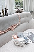 Balls of wool in basket on sofa, tablecloth with pink crocheted border over backrest