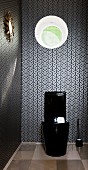 Black glossy toilet below round window in wall covered in black and white wallpaper with graphic pattern
