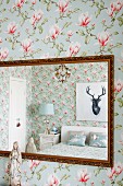 Portrait of stag above double bed reflected in mirror with antique gilt frame on magnolia-patterned floral wallpaper
