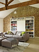 Modern grey sofa and matching ottoman in front of bookcase with cupboard element against brick wall