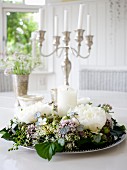 Wreath of flowers around white pillar candle on silver tray in front of silver candelabra