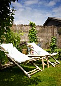 Bamboo sun loungers with pale cushions on lawn in front of willow screen