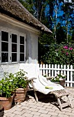 Holzliege mit weisser Decke, und Blumentöpfe auf Terrasse vor schlichtem Landhaus, in Sommerstimmung