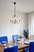 Crystal chandelier above set table with chairs upholstered in royal blue and vase of delphiniums in background