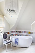 Ablage mit Mosaik aus bunten Scherben und Fliesenbordüre hinter freistehender Badewanne und einem Antikstuhl; Kronleuchter mit Schirm im Vordergrund