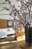 Half-height wood cladding behind white sofa combination, partitions made of bundled brushwood and vase of cherry blossom in foreground