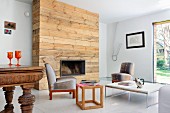 Rustic, untreated wood cladding on chimney breast in eclectic room with retro, upholstered easy chairs and various tables
